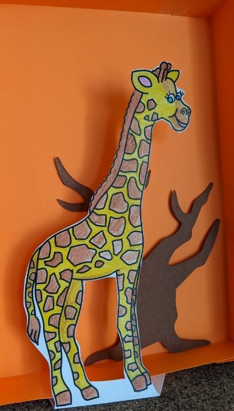 Giraffe paper cut-out in front of a paper box with a tree cut-out inside. The giraffe cut-out is casting a shadow into the paper box.