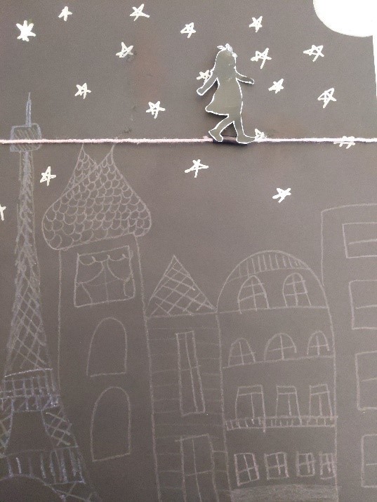 Craft made of paper and yarn, with a drawn-on background that depicts skyscrapers, stars, and a moon. A silhouette of a girl is attached to the yarn, which is pulled taut across the paper to resemble a high wire.