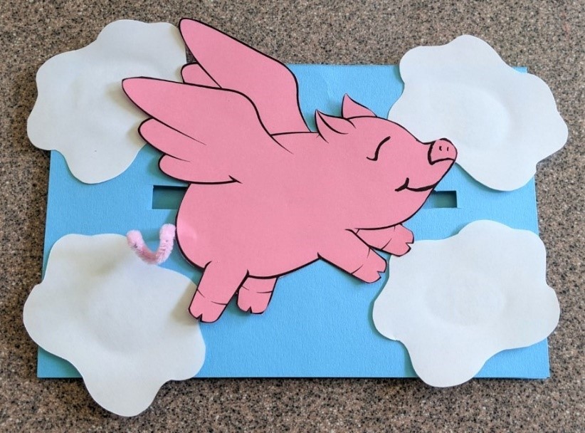Paper cut-out of a flying pig on top of a background made to look like the sky with paper cut-outs of clouds.