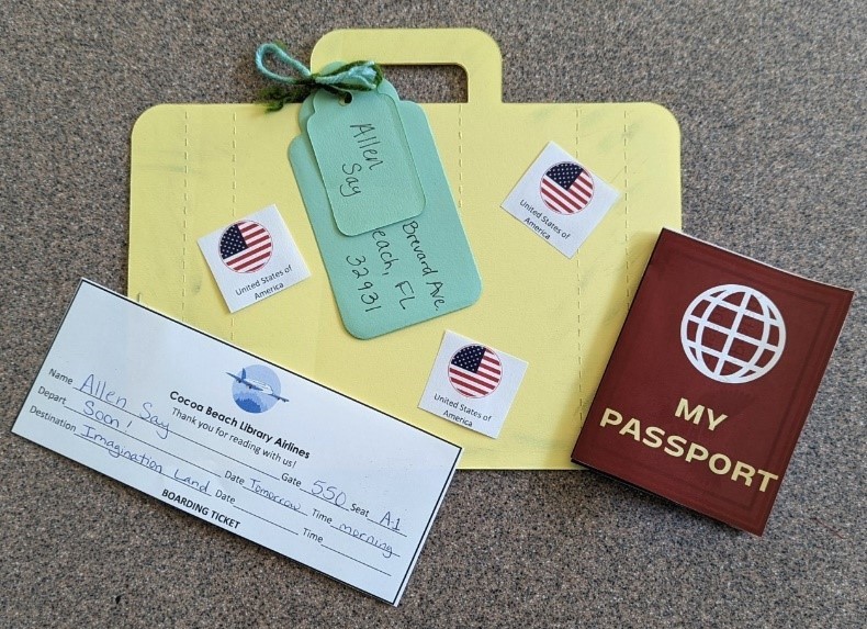 A large 2D paper suitcase sits below a play passport and Cocoa Beach Library Airlines boarding pass. Luggage tags are tied to the paper suitcase with yarn.